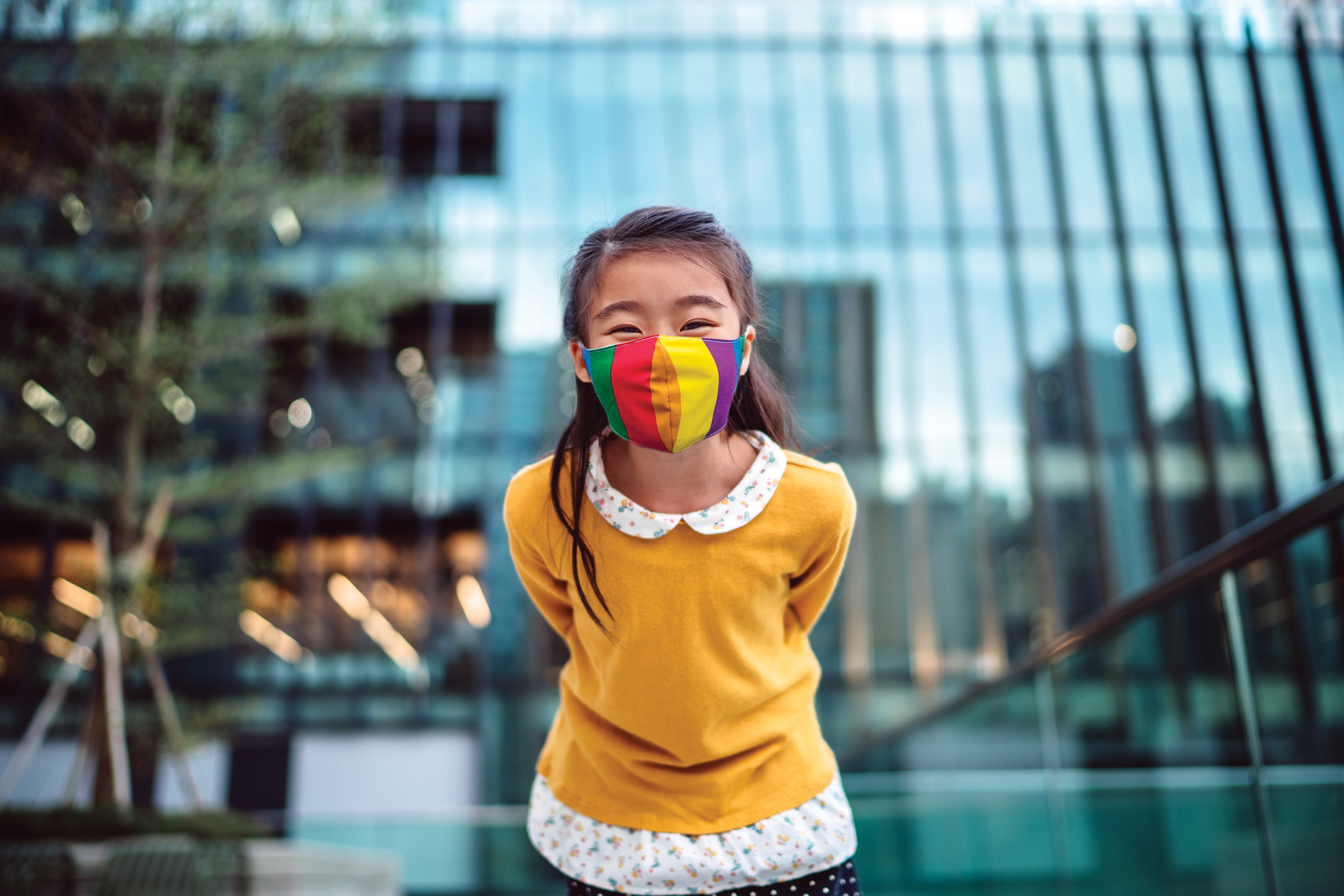 Une jeune fille portant un masque médical se penche vers la caméra et sourit joyeusement devant un immeuble de bureaux